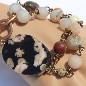 May include: A bracelet with a large, oval-shaped, black and white agate stone, surrounded by smaller white, brown, and red agate beads. The bracelet has a copper clasp.
