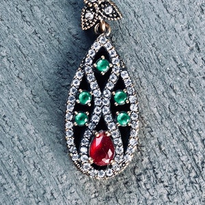 May include: A silver pendant with a teardrop-shaped design. The pendant is adorned with a red gemstone, green gemstones, and clear cubic zirconia stones.