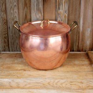 May include: A large copper pot with a lid and brass handles. The pot has a hammered finish and is sitting on a wooden surface.