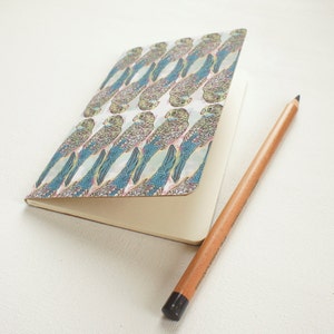 Puede incluir: Un cuaderno abierto con una cubierta que presenta un patrón repetido de ilustraciones de periquitos azules, verdes y beige. Un lápiz de madera con punta negra descansa junto al cuaderno abierto, que tiene páginas en blanco de color crema. El cuaderno está sobre una superficie blanca.