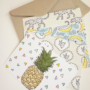 May include: A selection of illustrated greeting cards featuring a pineapple, bananas, and a leopard print design. The cards are stacked with a brown envelope. The pineapple card has colourful triangle accents.