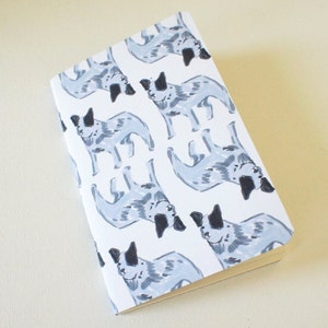 Puede incluir: Un cuaderno blanco con un patrón repetido de ilustraciones de perros azules y grises. Los perros tienen orejas y manchas negras, creando un diseño juguetón. El cuaderno tiene un borde de color crema claro.