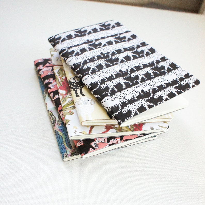 Puede incluir: Una pila de cinco cuadernos con cubiertas estampadas. El cuaderno superior tiene un estampado de guepardo en blanco y negro. Otras cubiertas presentan dise&ntilde;os florales e ilustrados en rosa, verde y azul. Los cuadernos tienen p&aacute;ginas blancas.