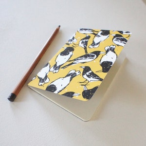 Puede incluir: Un pequeño cuaderno con una cubierta amarilla con un patrón de pájaros en blanco y negro. Un lápiz marrón descansa junto al cuaderno abierto. Las páginas del cuaderno son de color beige claro.
