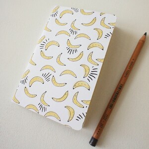 Puede incluir: Un cuaderno blanco con un patr&oacute;n repetido de pl&aacute;tanos amarillos y detalles en negro. Un l&aacute;piz marr&oacute;n con grafito negro descansa junto al cuaderno, listo para escribir o dibujar.