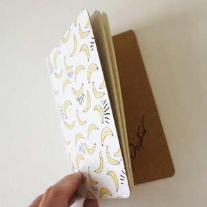 Puede incluir: Un cuaderno con una cubierta blanca con un patr&oacute;n de pl&aacute;tanos amarillos con detalles en negro. El cuaderno est&aacute; abierto, revelando las p&aacute;ginas interiores. Un bloc de notas rectangular marr&oacute;n es visible detr&aacute;s del cuaderno.
