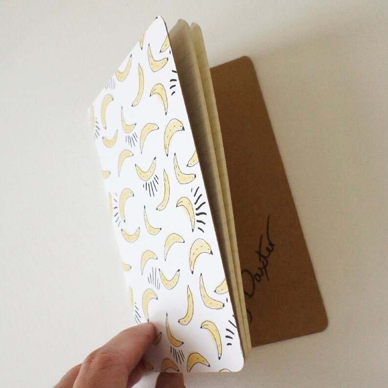 Puede incluir: Un cuaderno con una cubierta blanca con un patr&oacute;n de pl&aacute;tanos amarillos con detalles en negro. El cuaderno est&aacute; abierto, revelando las p&aacute;ginas interiores. Un bloc de notas rectangular marr&oacute;n es visible detr&aacute;s del cuaderno.