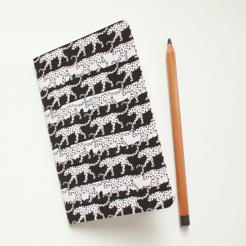 K&ouml;nnte beinhalten: Ein Notizbuch mit einem schwarz-wei&szlig;en Gepardenmuster. Ein Bleistift aus Holz liegt neben dem Notizbuch. Das Cover des Notizbuchs hat ein sich wiederholendes Muster von Geparden mit schwarzen Flecken.