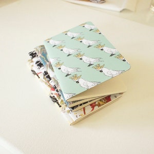 Puede incluir: Una pila de cuadernos peque&ntilde;os con cubiertas estampadas. El cuaderno superior tiene un fondo verde claro con cacat&uacute;as blancas. Otras cubiertas presentan dise&ntilde;os florales y abstractos en varios colores. Los cuadernos est&aacute;n apilados sobre una superficie blanca.