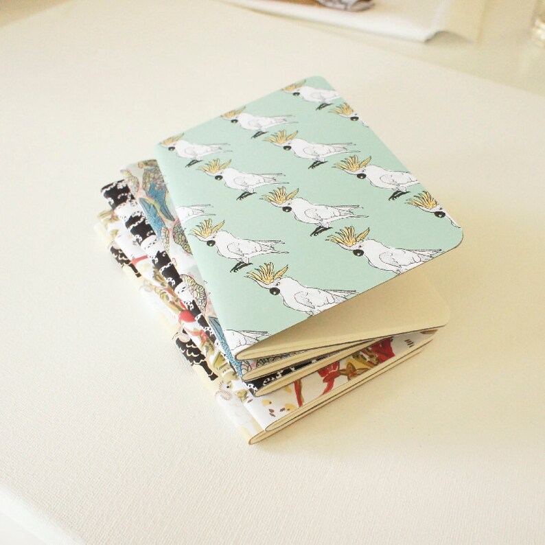 Puede incluir: Una pila de cuadernos peque&ntilde;os con cubiertas estampadas. El cuaderno superior tiene un fondo verde claro con cacat&uacute;as blancas. Otras cubiertas presentan dise&ntilde;os florales y abstractos en varios colores. Los cuadernos est&aacute;n apilados sobre una superficie blanca.