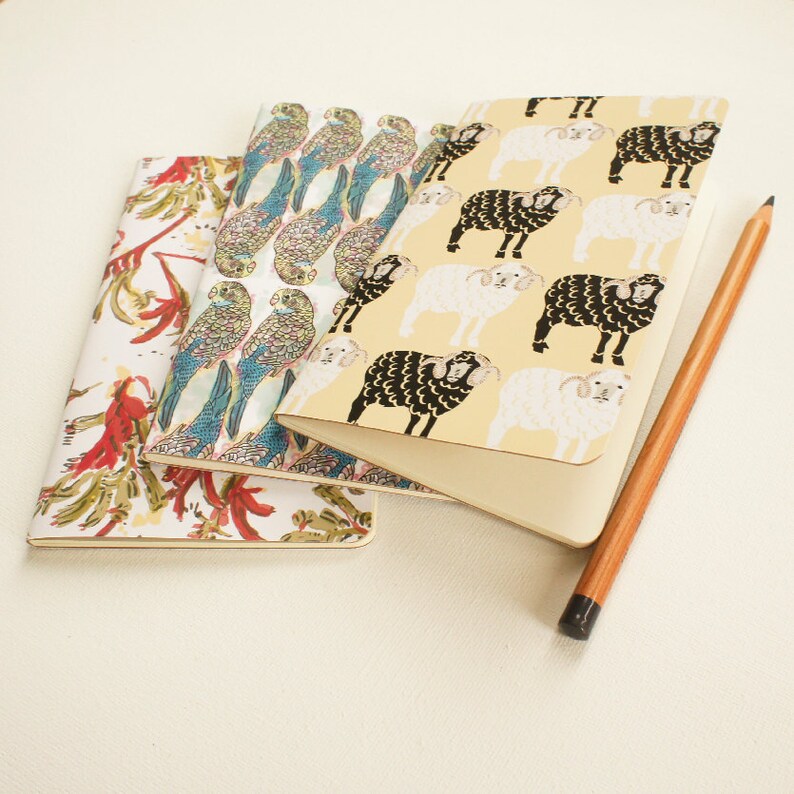 K&ouml;nnte beinhalten: Drei Notizb&uuml;cher mit farbenfrohen Einb&auml;nden und einem Bleistift. Die Notizb&uuml;cher zeigen Muster von Papageien, Schafen und floralen Designs in Rot, Wei&szlig; und Gelb. Der Bleistift ist braun mit einer schwarzen Spitze.