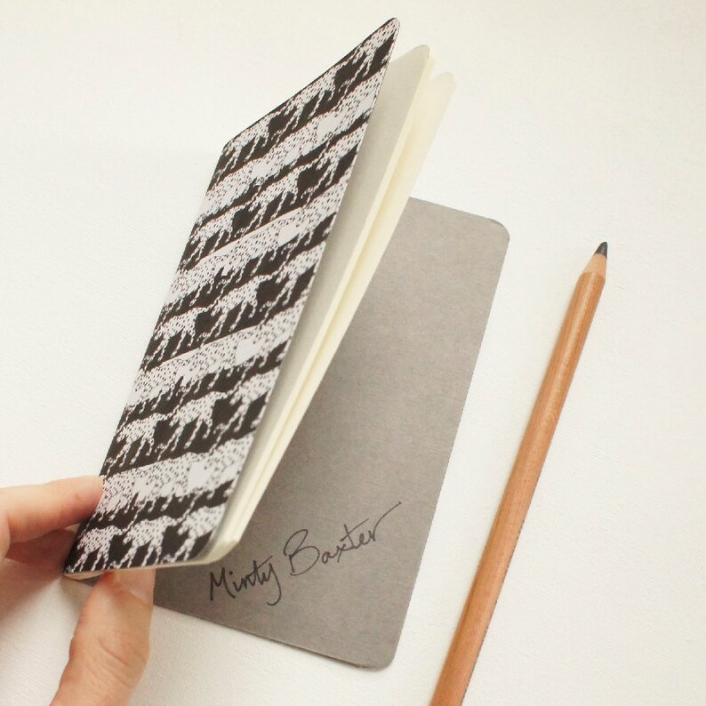 K&ouml;nnte beinhalten: Ein offenes Notizbuch mit einem schwarz-wei&szlig;en Muster auf dem Einband, gehalten von einer Hand. Die Innenseiten sind cremefarben. Ein Bleistift liegt neben dem Notizbuch. Der graue Einband tr&auml;gt die Signatur "Minty Baxter".