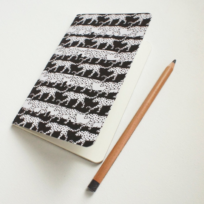 K&ouml;nnte beinhalten: Ein Notizbuch mit einem schwarz-wei&szlig;en Gepardenmuster auf dem Einband und leeren Seiten. Ein Bleistift aus Holz mit schwarzer Spitze liegt neben dem Notizbuch auf einer wei&szlig;en Oberfl&auml;che. Das Notizbuch ist ge&ouml;ffnet und zeigt die leeren Seiten.
