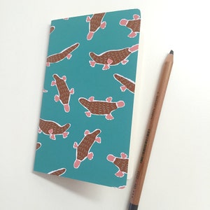 Puede incluir: Un cuaderno turquesa con un patrón repetido de ilustraciones de ornitorrincos marrones y rosas. Un lápiz de madera descansa junto al cuaderno. El cuaderno está cerrado y tiene una cubierta blanda.