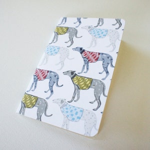 Puede incluir: Un cuaderno rectangular con fondo blanco que presenta un patrón repetido de galgos con suéteres de varios colores. Los suéteres son rojos, azules y amarillos, con un patrón de puntos en los perros.
