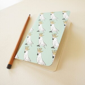 Puede incluir: Un pequeño cuaderno con una cubierta verde claro que presenta un patrón repetido de ilustraciones de cacatúas blancas con crestas amarillas. Un lápiz de madera con punta negra descansa junto al cuaderno sobre una superficie color crema.