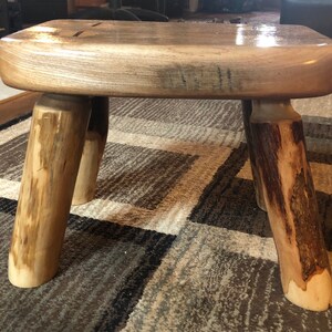 Peut inclure: Un tabouret en bois rustique avec une finition en bois naturel et quatre pieds. Le tabouret est fait de rondins bruts et a un design simple et artisanal.