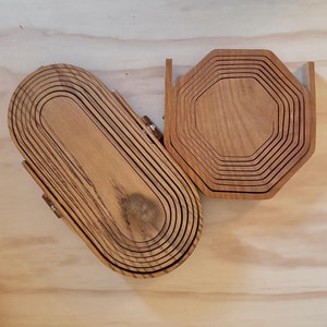 May include: Two wooden coasters, one oval and one octagonal, are stacked in concentric rings. The coasters are made of light brown wood with a natural grain pattern.