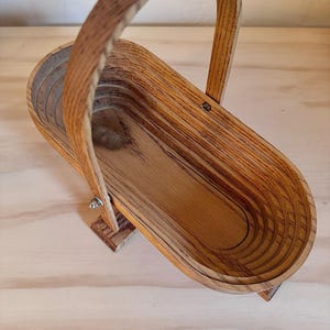 May include: A wooden basket with a handle, made of light brown wood with a natural grain pattern. The basket is folded and has a rounded oval shape.