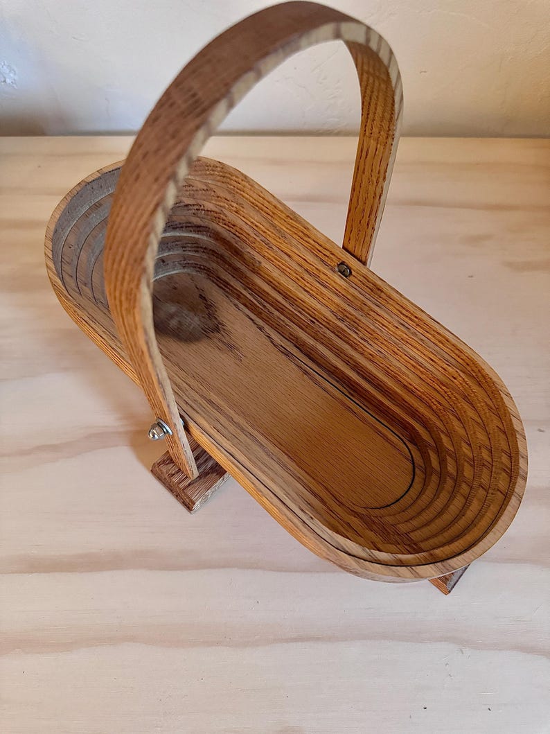 May include: A wooden basket with a handle, made of light brown wood with a natural grain pattern. The basket is folded and has a rounded oval shape.