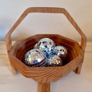May include: A wooden octagonal basket with a handle, sitting on a light wood surface. The basket is filled with silver disco balls.