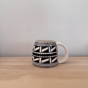 May include: A ceramic mug featuring a black and white geometric pattern. The design consists of horizontal bands, repeating diagonal rectangles, and a dotted line motif. The mug has a rounded body shape and a curved handle on the right side.