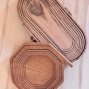 May include: Two wooden serving trays, one oval and one octagonal, are stacked on a wooden surface. Both trays are made of multiple layers of wood, creating a unique and rustic look.