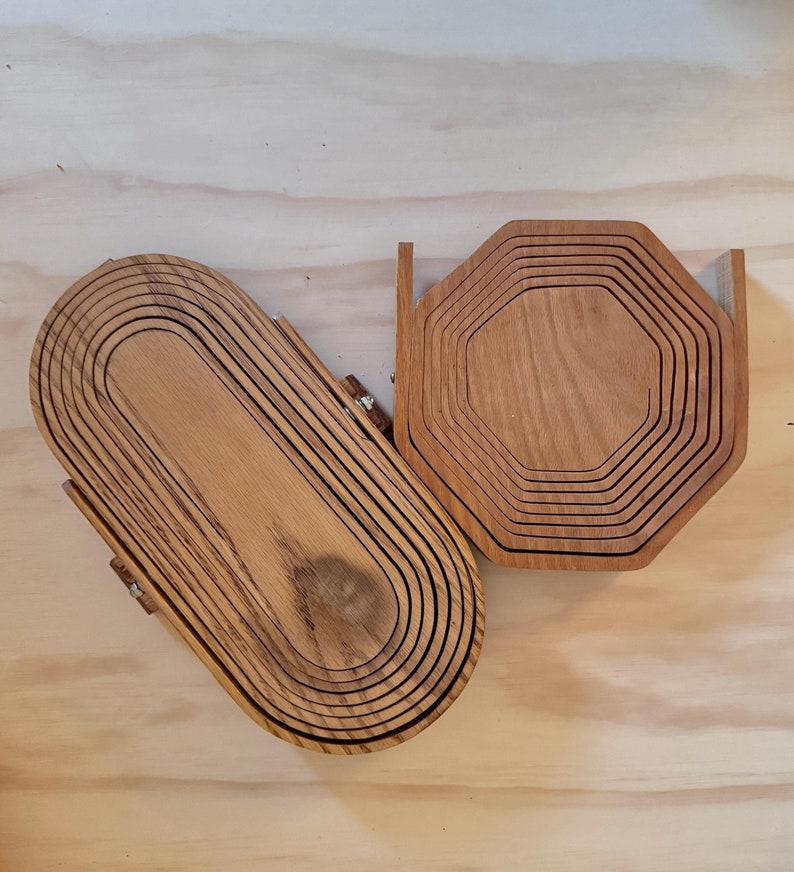 May include: Two wooden coasters, one oval and one octagonal, are stacked in concentric rings. The coasters are made of light brown wood with a natural grain pattern.