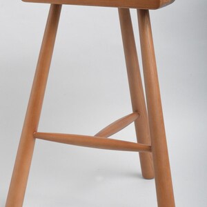 Könnte beinhalten: Ein Holzhocker mit einem sattelförmigen Sitz und drei Beinen. Der Hocker ist aus hellbraunem Holz gefertigt und hat ein schlichtes, rustikales Design.