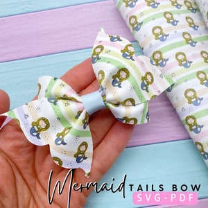 May include: A white bow with a mermaid tail pattern. The bow is made of fabric and has a light blue center. The fabric has a rainbow stripe pattern with stars and hearts. The text "Mermaid Tails Bow SVG-PDF" is at the bottom of the image.