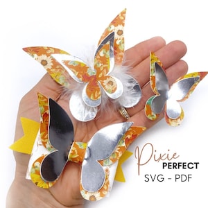 May include: Three paper butterflies with a floral pattern and silver accents. The butterflies are layered and have a 3D effect. The butterflies are on a hand with a yellow bow.
