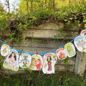 Könnte beinhalten: Eine Girlande aus bunten Papptellern mit Illustrationen von Menschen und Naturszenen. Die Teller sind an einem blauen Seil aufgehängt und mit Blumen, Bäumen und einem Lagerfeuer verziert. Der Text auf den Tellern lautet "Frühlingsfest".
