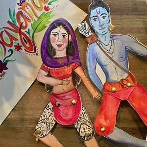 May include: Hand-painted paper cutouts of a man and woman in traditional Indian attire. The man, with blue skin, holds a bow and arrow. The woman wears a purple head covering and orange top. A watercolor painting with the word "Namaste" is in the background.