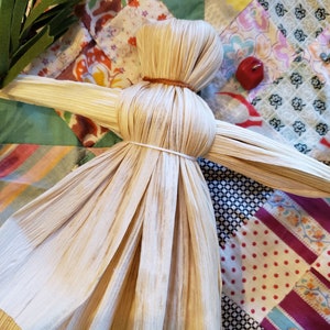 May include: A handmade corn husk doll with outstretched arms, lying on a colorful patchwork quilt.