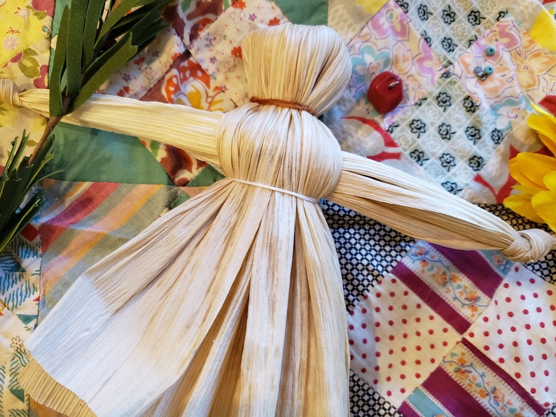 May include: A handmade corn husk doll with outstretched arms, lying on a colorful patchwork quilt.