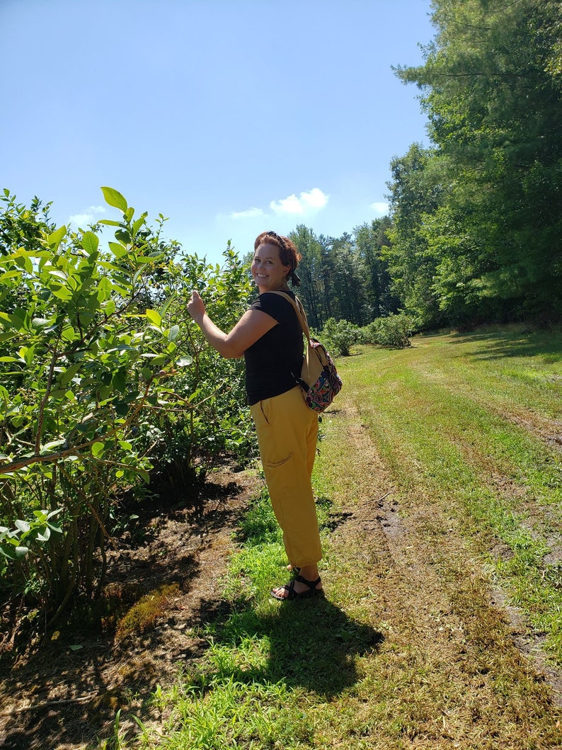 May include: A woman wearing a black shirt and yellow pants stands in a field of blueberry bushes. She is smiling and looking at the camera. She is holding a branch of blueberry bushes in her hand.