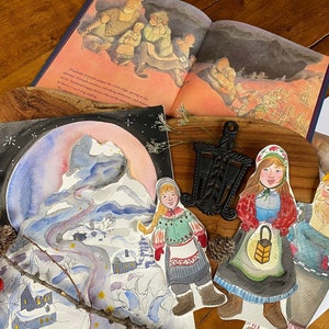 May include: A watercolor painting of a snowy mountain scene with a village at the base. The painting is surrounded by paper dolls of people in traditional clothing, including a woman holding a lantern and a man with a beard. The dolls are on a wooden surface with a book and a metal trivet.