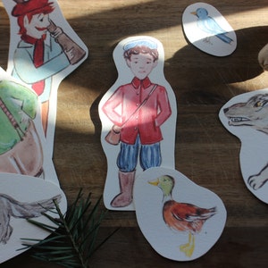 May include: A set of paper cutouts featuring characters from the fairy tale Little Red Riding Hood. The characters include Little Red Riding Hood, the wolf, the grandmother, the woodsman, and a bird. The cutouts are on a wooden surface.