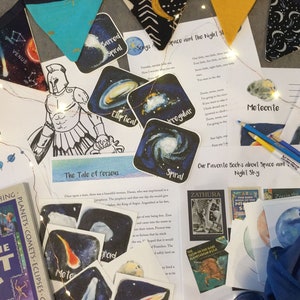 Peut inclure: Une collection de matériel éducatif sur le thème de l'espace, comprenant un livre intitulé "The Kids Book of the Night Sky", une feuille de travail imprimable intitulée "The Tale of Perseus" et plusieurs autocollants colorés sur le thème des galaxies. Le matériel est disposé sur un fond en tissu bleu et blanc avec des guirlandes lumineuses.