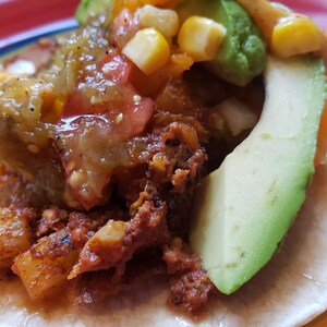 May include: A close-up of a taco filled with chorizo, potatoes, corn, tomatoes, avocado, and salsa verde. The taco is on a colorful plate.