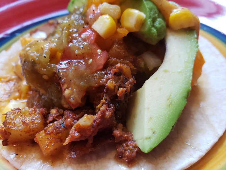 May include: A close-up of a taco filled with chorizo, potatoes, corn, tomatoes, avocado, and salsa verde. The taco is on a colorful plate.