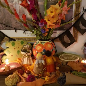 May include: A fall-themed altar with a variety of items, including a ceramic vase with orange and yellow flowers, a wooden leaf-shaped plate, a small woven basket, a lit candle, and a small doll. The altar is decorated with fall colors, such as orange, yellow, and brown.