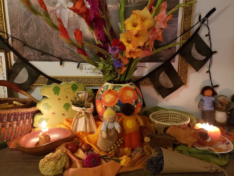 May include: A fall-themed altar with a variety of items, including a ceramic vase with orange and yellow flowers, a wooden leaf-shaped plate, a small woven basket, a lit candle, and a small doll. The altar is decorated with fall colors, such as orange, yellow, and brown.