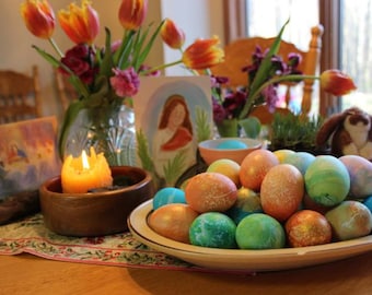 Pode incluir: Uma mesa posta com um vaso de tulipas laranja, uma vela acesa e uma tigela de ovos de Páscoa coloridos. Os ovos são pintados em tons de azul, verde e amarelo.