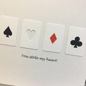 May include: A white greeting card featuring four playing card symbols: a black spade, a heart, a red diamond, and a black club. The phrase "You stole my heart" is printed below the cards.