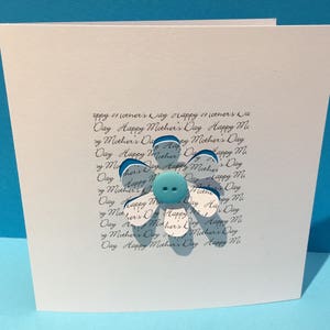 May include: A white card with a blue and white paper flower with a blue button in the centre. The card has the text "Happy Mother's Day" written on it multiple times.