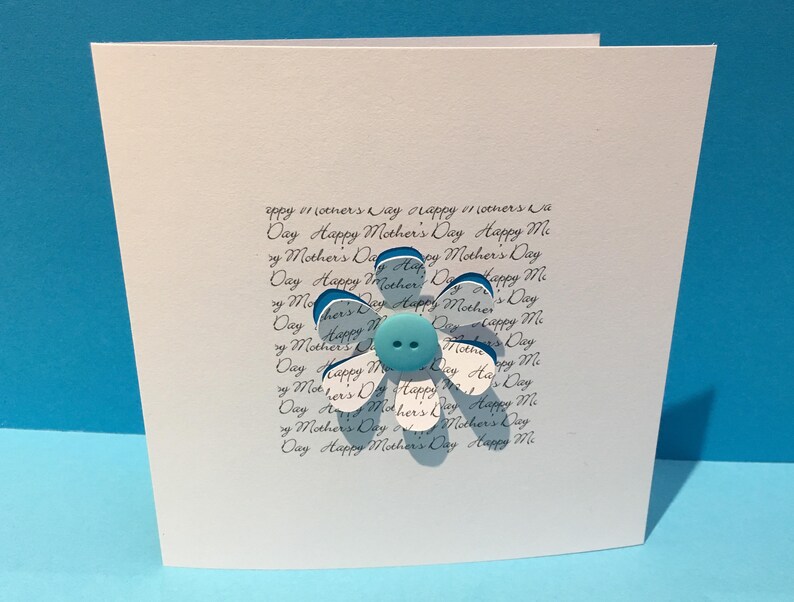 May include: A white card with a blue and white paper flower with a blue button in the centre. The card has the text "Happy Mother's Day" written on it multiple times.