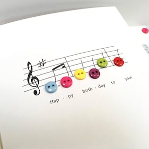 Può includere: Un biglietto di auguri bianco con un pentagramma musicale e bottoni colorati che compongono la frase "Happy birthday to you!". I bottoni sono blu, rosa, gialli, viola, verdi e rossi. Ulteriori bottoni sono sparsi.