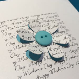 May include: A white card with a blue button in the centre of a paper flower. The flower is made of white paper with the words "Happy Mother's Day" repeated on each petal.
