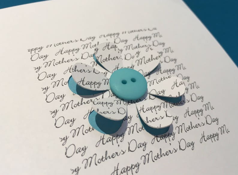 May include: A white card with a blue button in the centre of a paper flower. The flower is made of white paper with the words "Happy Mother's Day" repeated on each petal.